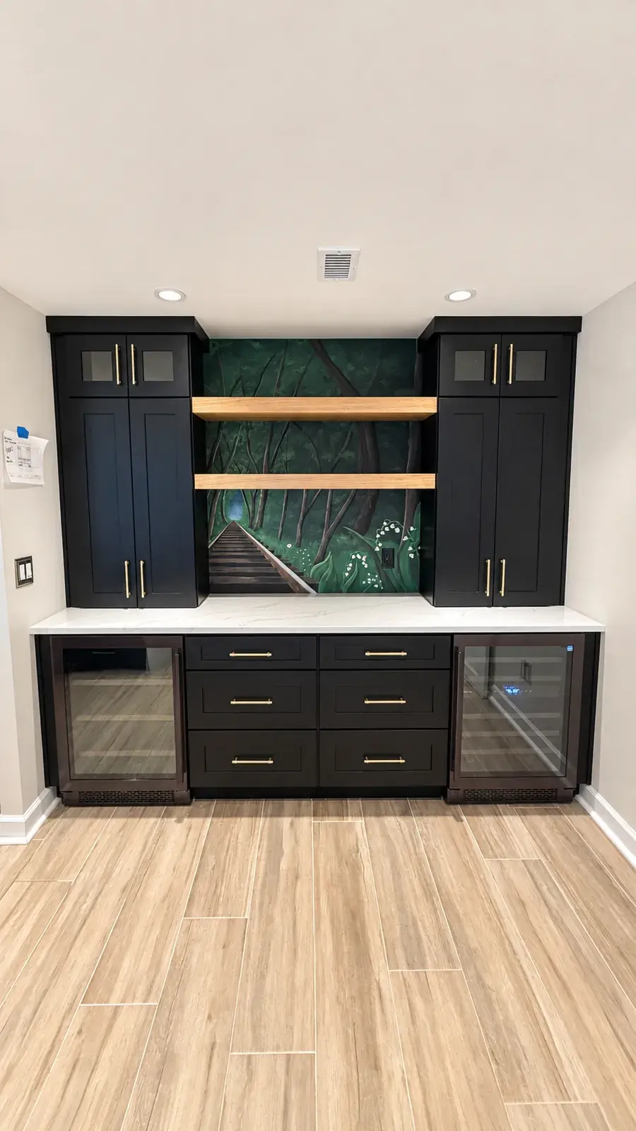 Built-in wet bar with black shaker cabinets, two wine refrigerators, oak floating shelves, and a forest mural