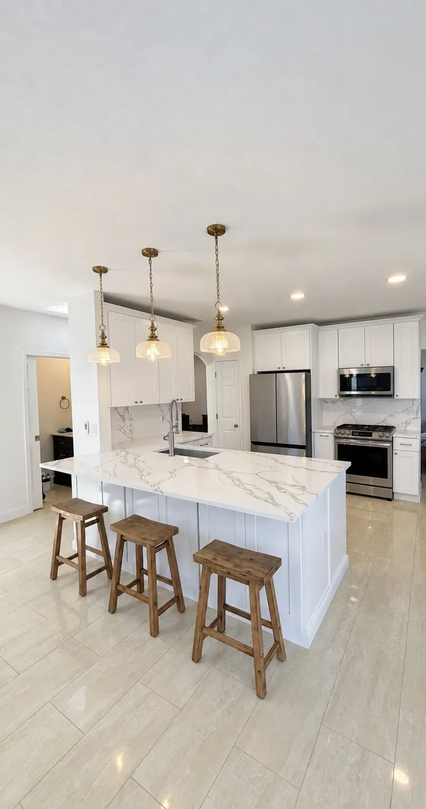 White shaker kitchen with a marble-look quartz island, three wood stools, and brass pendant lights