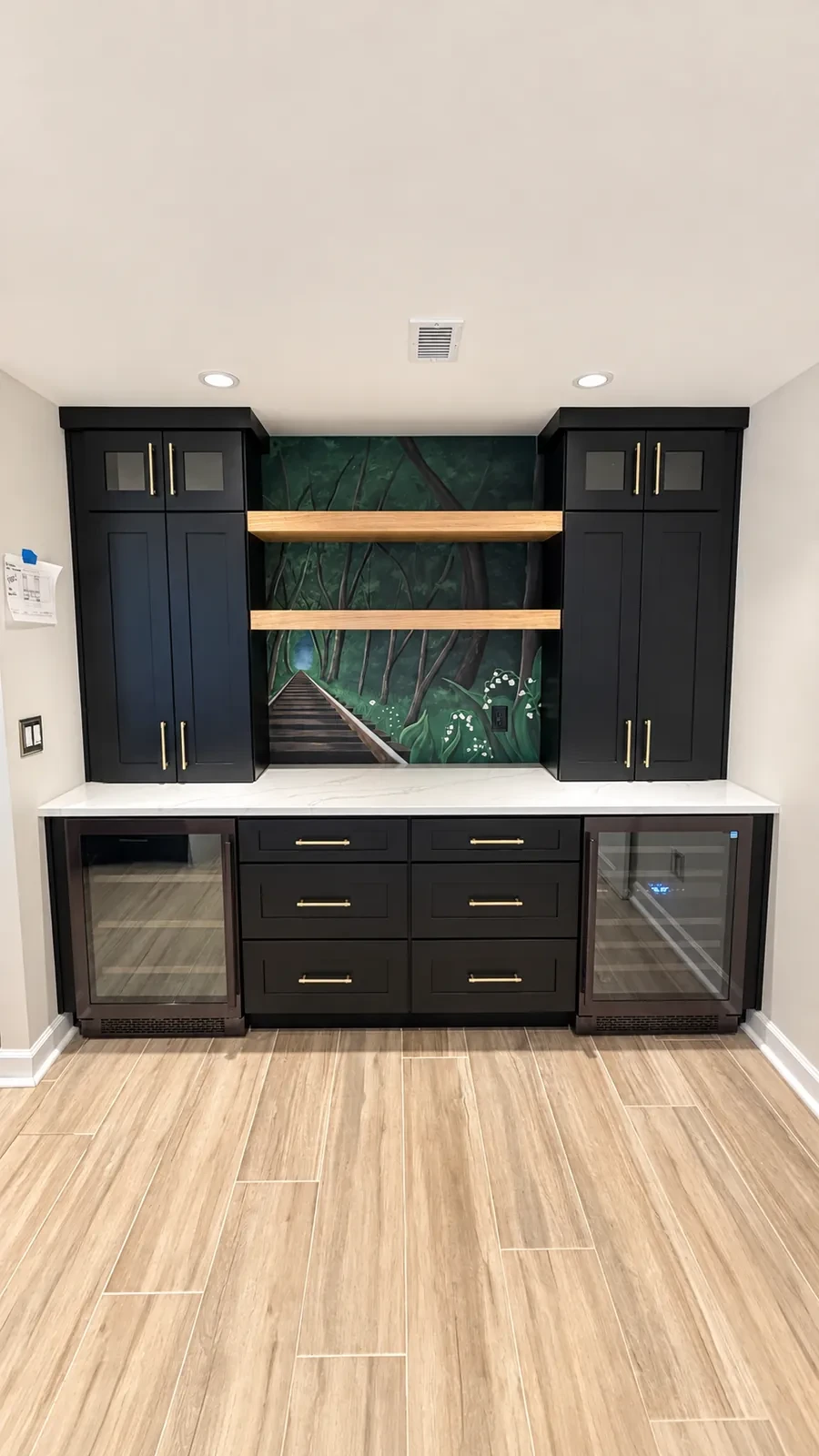 Built-in wet bar with black shaker cabinets, two wine refrigerators, oak floating shelves, and a forest mural