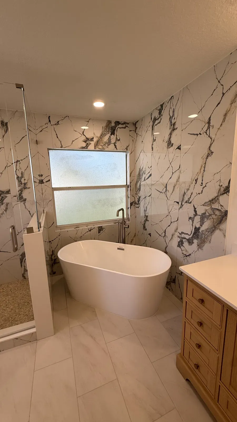 Marble-look tile shower with a glass panel and a freestanding tub under a frosted window