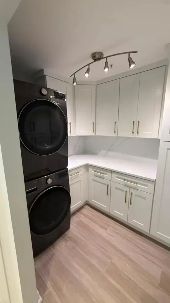 Stacked washer and dryer beside white cabinets with brass pulls and track lighting Stacked washer and dryer beside white cabinets with brass pulls and track lighting