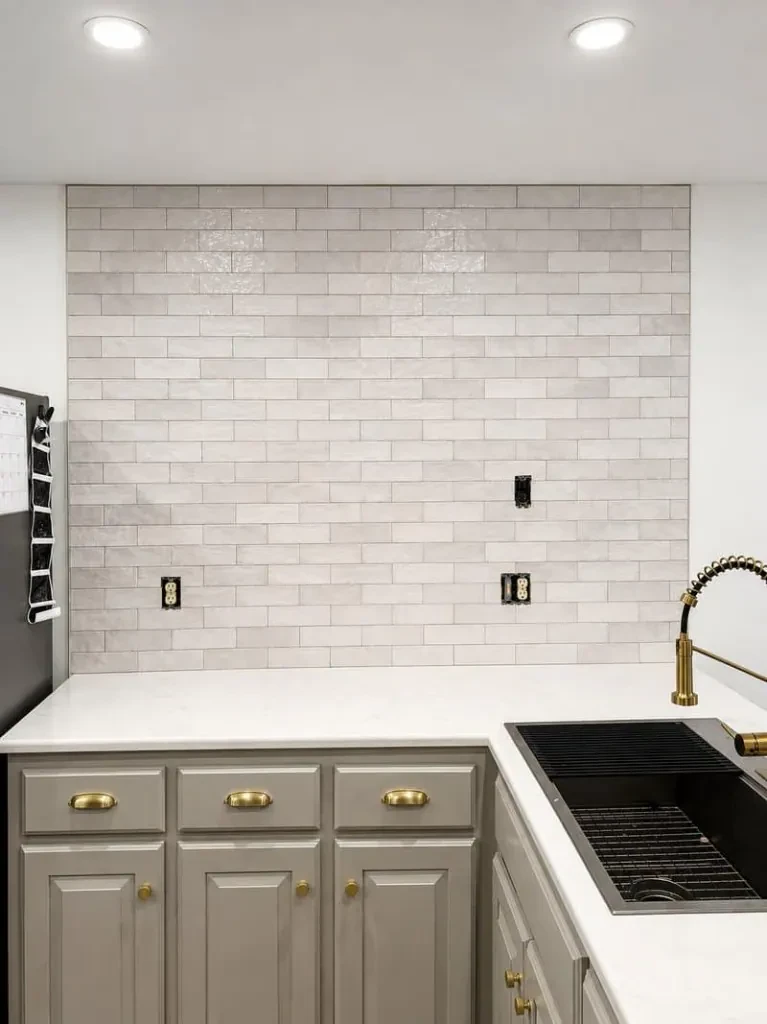 Glossy white subway-tile backsplash above a white counter, gray cabinets, and a brass faucet